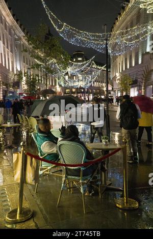 UK Weather, Londra, 12 novembre 2023: Le luci di Natale su Regent Street si riflettono sulle strade bagnate in una notte piovosa. Nonostante il tempo la gente si siede fuori da un caffè a un tavolo, riparandosi sotto un ombrello. Anna Watson/Alamy Live News Foto Stock
