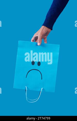 primo piano di un uomo che tiene in mano un sacchetto di carta blu vuoto, con una faccia triste dipinta al suo interno, capovolta su uno sfondo blu Foto Stock