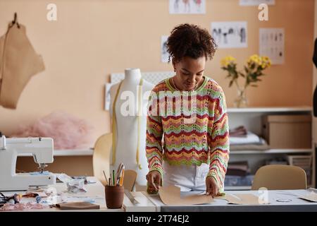 Giovane artigiana creativa che si piega sopra il tavolo con motivi di cucito e tessuti mentre lavora su nuovi capi di abbigliamento nella sua officina Foto Stock