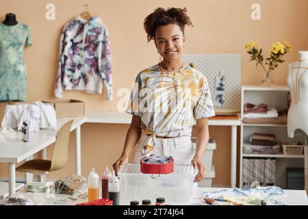 Felice giovane designer afroamericana con t-shirt tie dye in piedi accanto al posto di lavoro con bottiglie di vernici coloranti e altre forniture Foto Stock