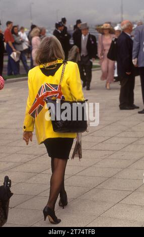 Elegante donna inglese alla moda anni '1990 vestita da una giacca gialla brillante e da Una gonna nera alla moda A line, collant neri e scarpe nere. Due bandiere Union Jack sono nella sua borsa nera. Derby Day corse di cavalli Epsom Downs, Surrey, Inghilterra giugno 1990 1990s UK HOMER SYKES Foto Stock