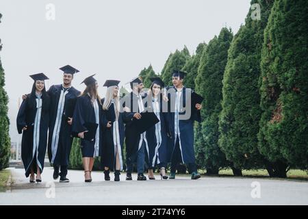 Diversi diplomati universitari indossano abiti per la laurea che celebrano il successo con gli amici e i docenti in un parco soleggiato. Foto Stock