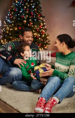 Foto verticale di una felice donna afroamericana che regala un regalo a suo figlio e marito, Natale Foto Stock