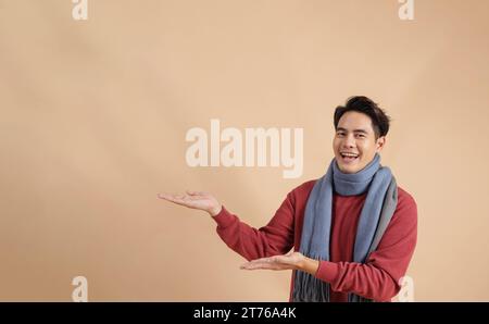Uomo asiatico felice e positivo in maglione rosso e sciarpa puntando le dita ad uno spazio vuoto isolato su sfondo di colore beige. Foto Stock