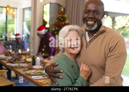 Due amici anziani felici e diversificati, uomini e donne, che si abbracciano, a pranzo di natale con gli amici Foto Stock