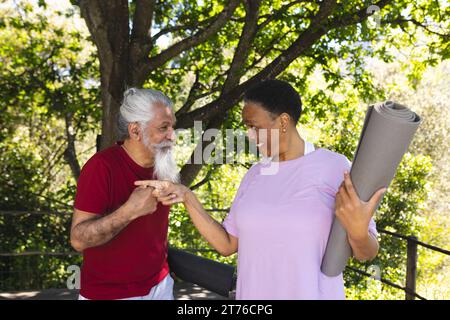 Due amici anziani felici e diversificati che parlano e sorridono alla lezione di yoga nel giardino soleggiato Foto Stock