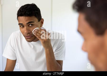 Uomo birazziale felice che pulisce il viso con un batuffolo di cotone che guarda nello specchio in bagno a casa Foto Stock