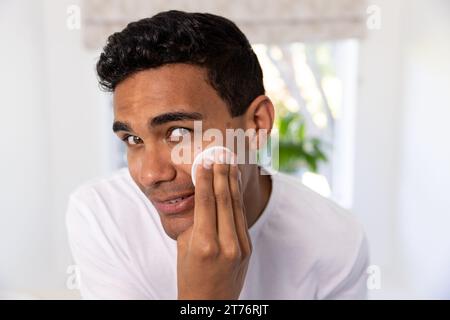 Un uomo birazziale felice che pulisce il viso con un batuffolo di cotone in bagno a casa Foto Stock