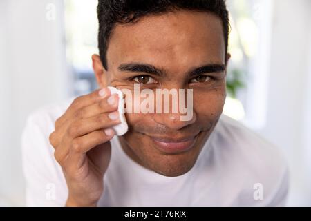 Un uomo birazziale felice che pulisce il viso con un batuffolo di cotone in bagno a casa Foto Stock