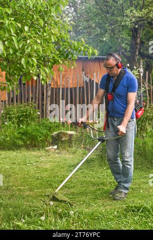 Un lavoratore taglia l'erba sul prato in giardino con un rasaerba portatile. Foto Stock