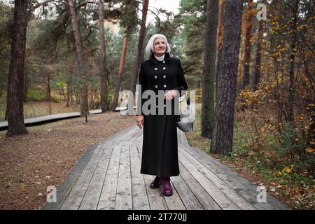 Graziosa donna matura vestita con elegante cappotto nero, passeggia attraverso l'incantevole foresta, immergendoti nello splendore della natura con un sorriso sereno, grazia e relax Foto Stock