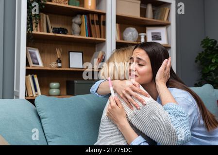 Una giovane donna felicissima che tiene un test di gravidanza dice a sua madre che è incinta e che avrà un bambino. Futura nonna e madre felice a casa si Foto Stock