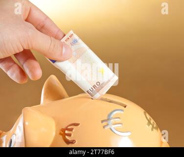 dettaglio di una mano che inserisce una banconota da cinquanta euro in un salvadanaio, concetto di business e risparmio su sfondo dorato Foto Stock