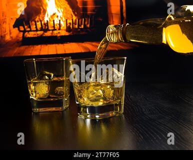 uomo che versa due bicchieri di whisky con cubetti di ghiaccio davanti al caminetto su un tavolo di legno Foto Stock