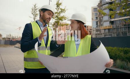 Caucasico due persone colleghi colleghi costruttori architetti ingegneri donna e uomo in caschi elmetti elmetti e giubbotti discutono di un documento cartaceo Foto Stock