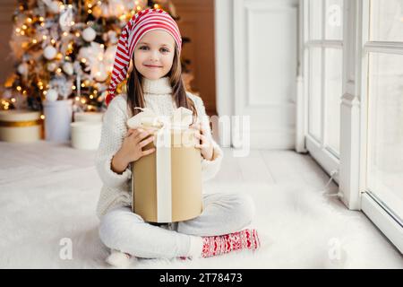 Bambino gioioso in abiti festivi con un grande regalo di Natale, con dietro un albero splendidamente adornato Foto Stock