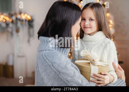 Un momento commovente in cui una madre abbraccia sua figlia, che tiene un regalo, con luci festive sullo sfondo Foto Stock