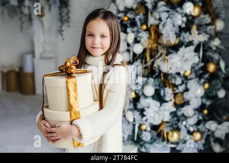 Una giovane ragazza sorridente che regge un regalo splendidamente avvolto davanti a un albero di Natale natalizio ornato d'oro Foto Stock