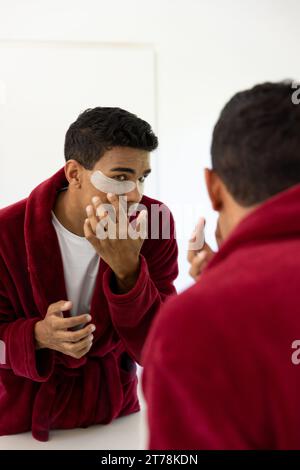 Uomo birazziale che applica cerotti sotto gli occhi che guardano nello specchio in bagno a casa Foto Stock