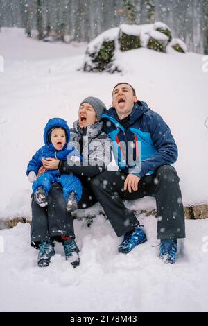 Mamma, papà e una bambina sono seduti su un ceppo nella foresta sotto una nevicata e prendono fiocchi di neve in bocca Foto Stock