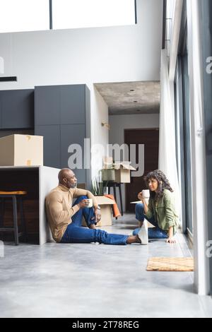 Coppia felice e diversificata seduta sul pavimento a bere caffè vicino alle scatole nella nuova casa, spazio fotocopie Foto Stock