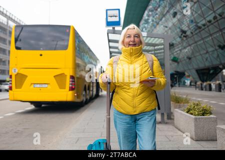 Felice viaggiatrice anziana in un'elegante giacca gialla e jeans con valigia, passaporto e biglietti presso una fermata dell'autobus in attesa di un autobus Foto Stock