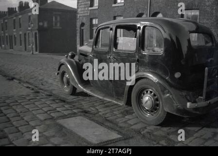 Anni '1950, storica, un'automobile di un decennio precedente, probabilmente degli anni '1940, parcheggiata su una strada acciottolata di piccole case vittoriane terrazzate, Oldham, Manchester, Inghilterra, Regno Unito. Foto Stock