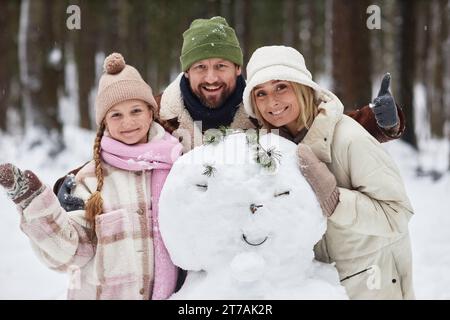 Allegra famiglia di tre persone in abbigliamento invernale in piedi accanto al pupazzo di neve di fronte alla macchina fotografica e che ti guarda mentre l'uomo barbuto mostra il pollice in alto Foto Stock
