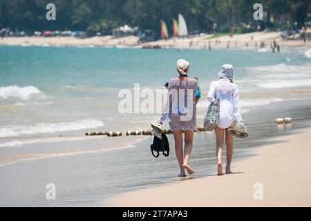 Due donne camminano sulla spiaggia di sabbia lungo l'acqua. Soleggiata giornata tropicale sulla spiaggia di Bang Thao, foto ravvicinata. PHUKET, THAILANDIA - 7 APRILE 2023 Foto Stock