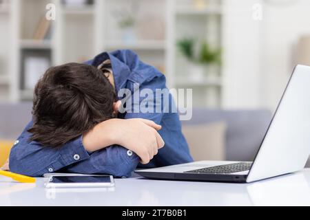 Studente stanco seduto alla scrivania di fronte al computer portatile durante la lezione a distanza, adolescente stanco e assonnato sdraiato sulle mani sulla scrivania con la testa abbassata, stanco di studiare. Foto Stock