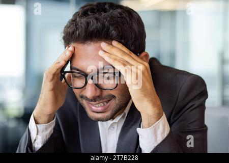 Ritratto ravvicinato di un uomo d'affari indiano con occhiali che soffre di mal di testa, seduto a una scrivania in un ufficio luminoso, che lavora, tenendo la testa con le mani. Foto Stock