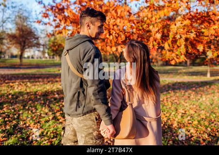 Vista posteriore del soldato in uniforme ucraina e di sua moglie che cammina nel parco autunnale. Coppia che si abbraccia dopo una lunga separazione. Ritorno a casa Foto Stock