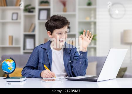 Un simpatico studente siede davanti a un notebook a un tavolo in una stanza luminosa di casa, studia in remoto, guarda lo schermo del computer, ondate di saluto, scrive in un notebook. Foto Stock