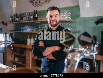 Ritratto del barman barbarbato sorridente vestito in uniforme nera con un grembiule al bancone del bar con rubinetti di birra alla spina. Persone di successo, duro lavoro, Foto Stock