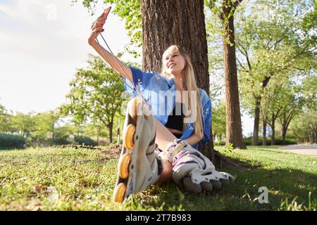 donna bionda seduta su un albero con pattini in linea mentre scatta un selfie con il suo smartphone Foto Stock