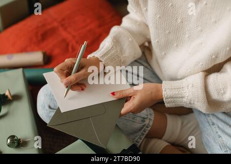 Primo piano di una donna seduta a gambe incrociate sul pavimento accanto a un regalo di Natale confezionato che scrive un biglietto di Natale Foto Stock