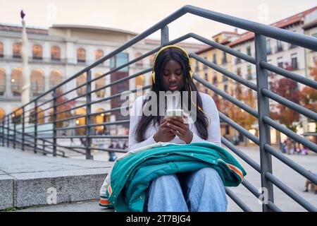 Ritratto di una giovane studentessa afroamericana all'interno di un campus universitario. Un giovane studente universitario nel campus siede su una scala usando p Foto Stock