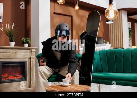 Il turista con cellulare e attrezzatura da snowboard nella hall dell'hotel si gusta una bevanda. Nella foto dell'attrezzatura invernale, il viaggiatore si rilassa vicino al caminetto nell'area lounge della stazione sciistica. Ideale per le vacanze invernali. Foto Stock