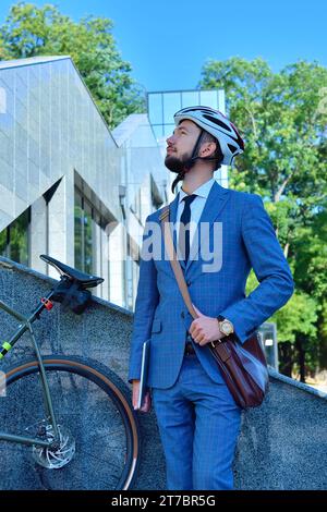 Uomo d'affari barbuto con casco e stand per biciclette vicino al business center. Stile business e urbano. Foto Stock