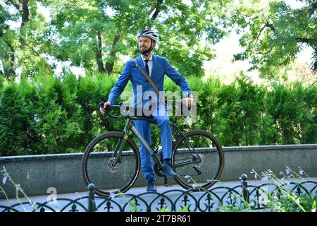 Uomo d'affari barbuto con casco e tuta con bicicletta. Stile business e urbano. Foto Stock