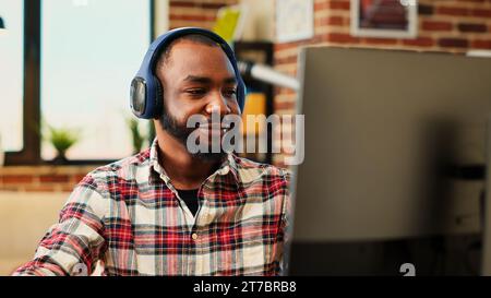 Lavoratore remoto felice che ascolta musica con le cuffie mentre lavora da casa. Rilassato e sorridente freelance che si diverte, scrivendo al computer nell'elegante ufficio personale dell'appartamento, da vicino Foto Stock