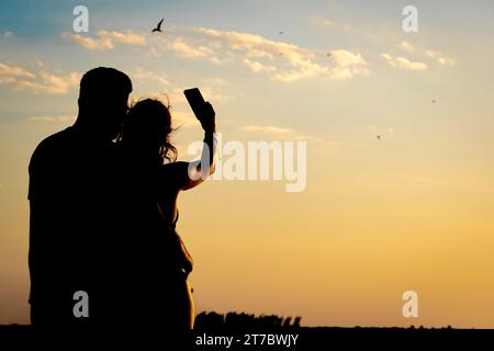Silhouette di una coppia che abbraccia, scattando una foto con il cellulare, gli uccelli che volano sul mare durante un tramonto bellissimo e romantico. Concetto o Foto Stock
