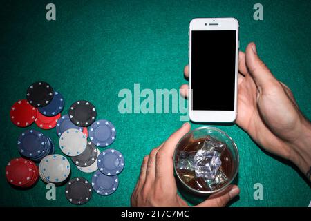 Mano del giocatore di poker che mostra il telefono cellulare che tiene il tavolo da poker in vetro del whiskey Foto Stock