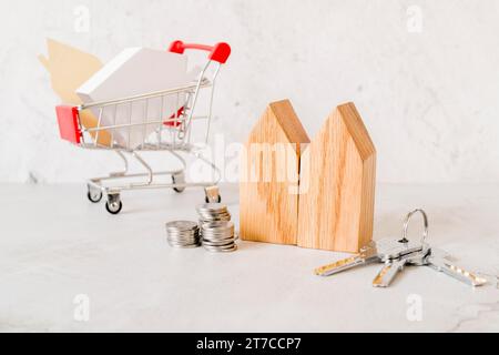 Blocchi di case in legno impilano monete, chiavi, piccolo carrello della spesa, sfondo in cemento Foto Stock