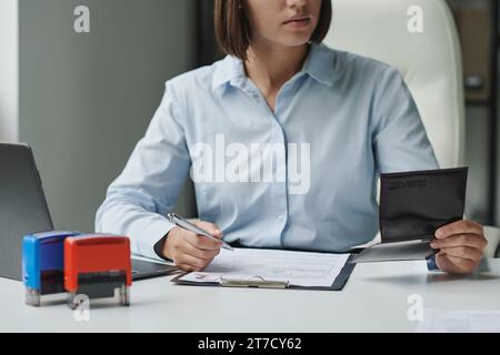 Giovane specialista che controlla il passaporto mentre compila i documenti per il visto sul posto di lavoro in ufficio Foto Stock