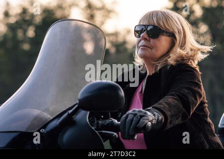 Bionda sicura e matura con guanti in pelle che guidano la sua moto il giorno autunnale. Turismo motociclistico hobby. Stile di vita sano. Sollievo emotivo. Excell Foto Stock