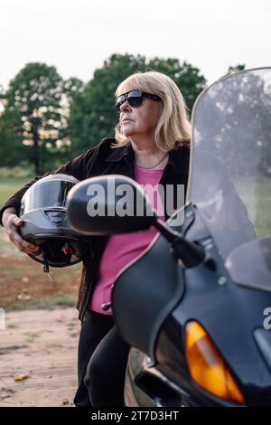 Ritratto verticale di bionda matura in giacca con casco in mano in piedi vicino alla moto in autunno in natura. Turismo motociclistico. Lifest sano Foto Stock