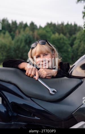 Ritratto verticale di una donna bionda matura con una chiave inglese sulla sua moto nera in natura il giorno d'autunno. Hobby turismo motociclistico.manutenzione motociclistica Foto Stock