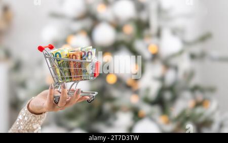 Un piccolo carrello pieno di banconote in euro tenute per mano di una donna con un albero di Natale sullo sfondo. Foto Stock