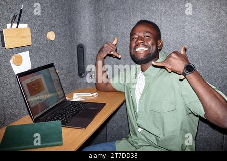 Uomo d'affari afro-americano felice seduto al suo posto di lavoro e lavorando online su un portatile Foto Stock